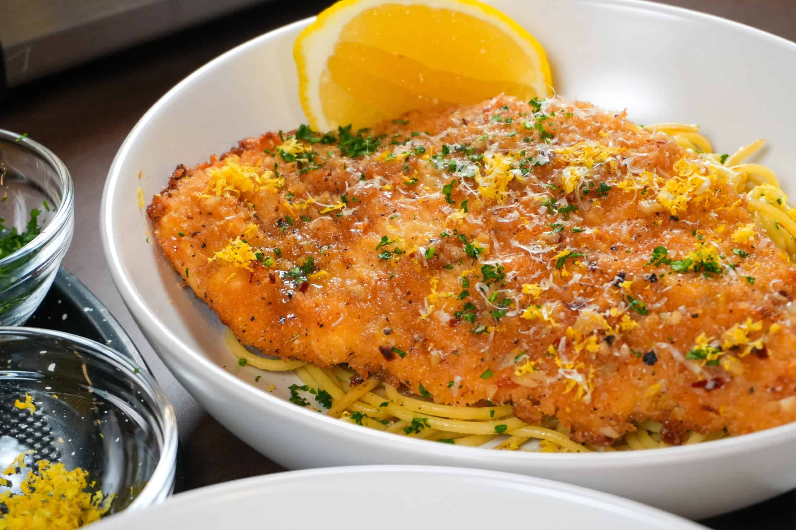 Crispy golden chicken cutlet on pasta with lemon and fresh herbs in a white bowl.